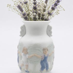 Peut inclure: Un vase en porcelaine blanche doté de deux anses en forme de volutes et d'un relief représentant deux enfants se tenant la main en courant parmi des arbres. Le vase contient un bouquet de brins de lavande violette et de fleurs de gypsophile.