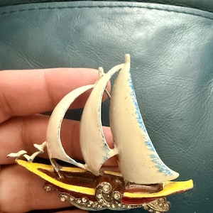 May include: Vintage sailboat brooch with cream sails and a yellow and brown hull. The brooch is decorated with rhinestones and has a pin closure. The sails have blue accents.