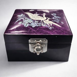 May include: A black lacquered box with a purple lid featuring a mother-of-pearl inlay design of two peacocks and floral elements. The box has a silver-colored clasp.