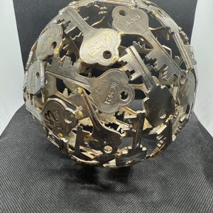Puede incluir: Una escultura esférica compuesta por numerosas llaves de metal, creando un diseño calado texturizado. Las llaves son de varios tamaños y formas, con marcas visibles. El color general es una mezcla de plata y oro, dándole una estética vintage.