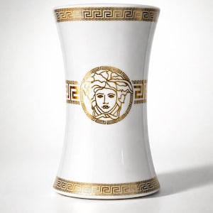 May include: White ceramic vase with gold Greek key pattern bands at the top and bottom, and a gold Medusa head emblem in the center. The vase has a slightly hourglass shape.