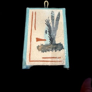 Op de afbeelding: Een kleine, rechthoekige hanger met een geschilderde illustratie van een vogel en een plant. Het kunstwerk heeft een lichtblauwe rand en een goudkleurige lus aan de bovenkant om op te hangen. De vogel is afgebeeld met een lange snavel en de plant heeft drie hoge bladeren.