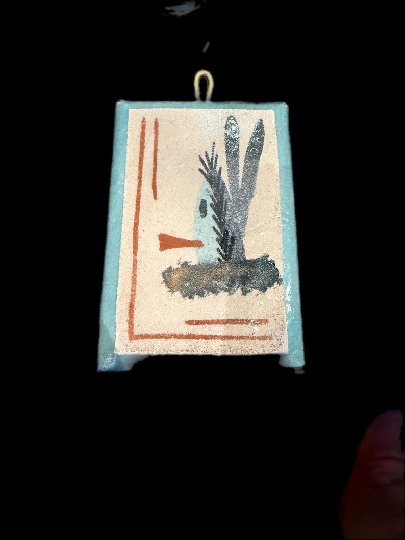 Op de afbeelding: Een kleine, rechthoekige hanger met een geschilderde illustratie van een vogel en een plant. Het kunstwerk heeft een lichtblauwe rand en een goudkleurige lus aan de bovenkant om op te hangen. De vogel is afgebeeld met een lange snavel en de plant heeft drie hoge bladeren.