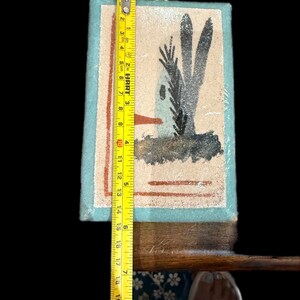 Op de afbeelding: Een ingelijst kunstwerk met een woestijnsc&egrave;ne met een huis en cactussen, ongeveer 20 cm hoog. Het heeft een lichtblauwe rand en een gouden ophanglus. De meting wordt gedaan met een gele meetlint.