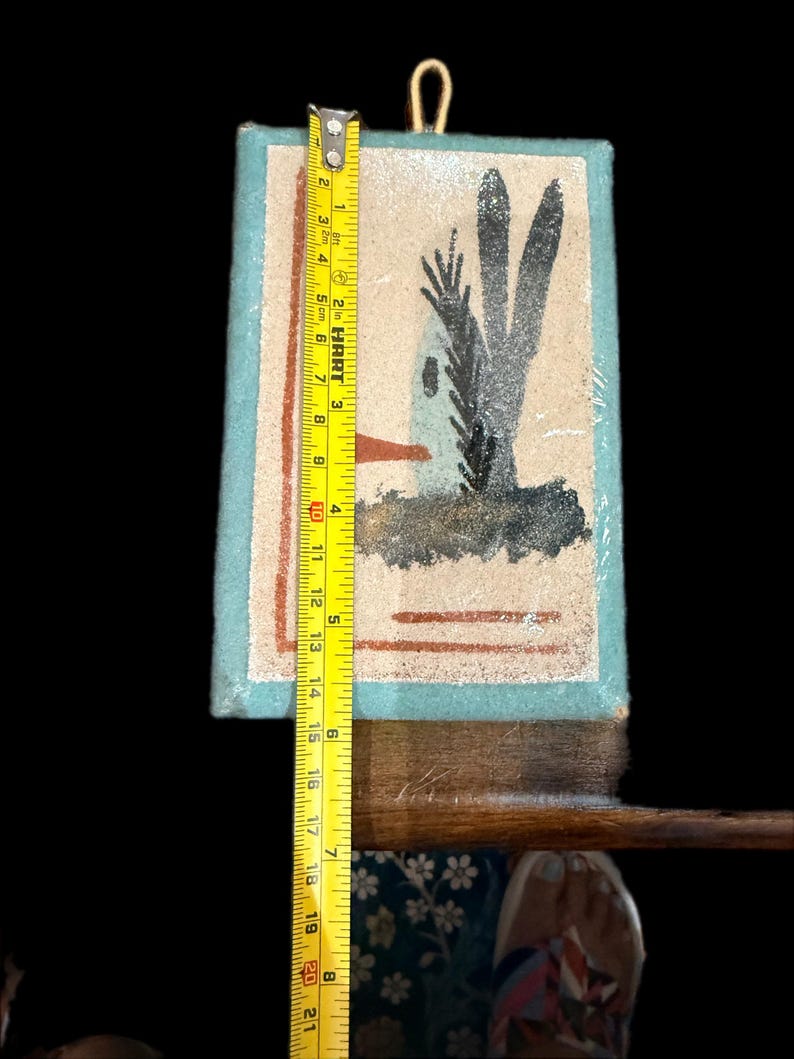 Op de afbeelding: Een ingelijst kunstwerk met een woestijnsc&egrave;ne met een huis en cactussen, ongeveer 20 cm hoog. Het heeft een lichtblauwe rand en een gouden ophanglus. De meting wordt gedaan met een gele meetlint.