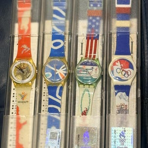 May include: Four Swatch Quartz watches in clear plastic cases. The watches have various strap designs, including Olympic and flag-themed patterns. The watch faces feature different designs and colors. The watches are labeled with the Swatch Quartz brand.