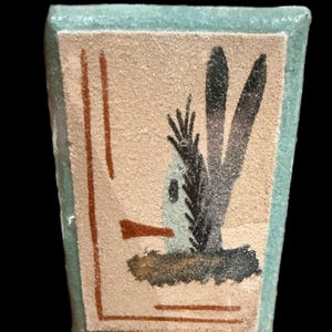 Op de afbeelding: Kleine, rechthoekige keramische tegel met een lichtbeige achtergrond en een lichtblauwe rand. De tegel toont een gestileerd woestijnlandschap met bruine en zwarte penseelstreken. Twee verticale bruine lijnen aan de linkerkant.