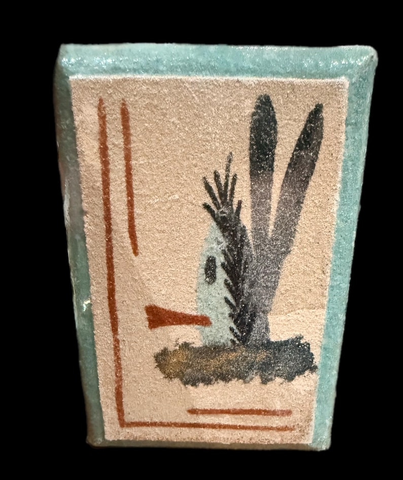 Op de afbeelding: Kleine, rechthoekige keramische tegel met een lichtbeige achtergrond en een lichtblauwe rand. De tegel toont een gestileerd woestijnlandschap met bruine en zwarte penseelstreken. Twee verticale bruine lijnen aan de linkerkant.