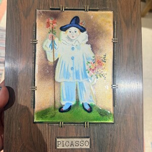 May include: A rectangular wooden wall plaque featuring a central enameled tile depicting a figure in a blue hat and white ruffled costume holding a flower. The tile is framed by metal clips and includes a small metal plate with the text PICASSO.