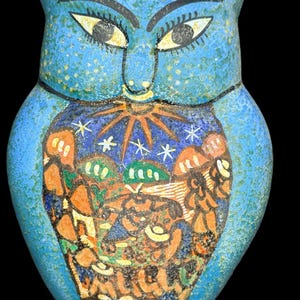May include: A blue owl-shaped wall hanging with a detailed, colorful design. The owl has large yellow eyes, black eyebrows, and a decorative belly featuring a landscape with a sun, stars, and trees. The owl is made of a textured material.