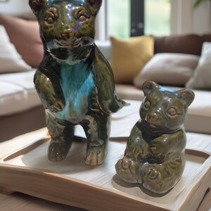May include: Two ceramic bear figurines with a glossy finish, one standing and one sitting, featuring dark olive green glaze with bright turquoise accents on the chest and face, displayed together on a light-colored rectangular wooden tray.