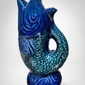 May include: A ceramic pitcher shaped like a fish with a glossy finish. The head is a deep royal blue, transitioning into a teal body with textured scales. The tail curves to form a handle, and the base is a solid dark blue with swirl patterns.