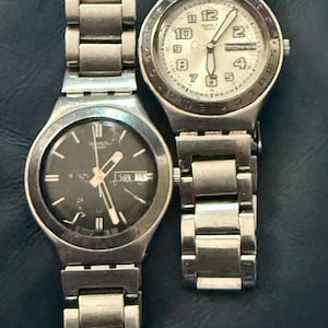 Puede incluir: Dos relojes Swatch con correas de metal plateado y esferas redondas. Uno tiene una esfera negra con manecillas y marcadores blancos, el otro tiene una esfera blanca. Ambos relojes muestran el día de la semana.