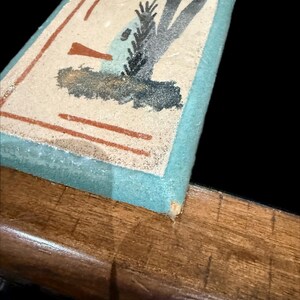 Op de afbeelding: Een rechthoekige wanddecoratie met een lichtblauwe rand en een beige midden. Het midden heeft een abstract ontwerp met bruine, zwarte en blauwe vormen. Er is een kleine goudkleurige haak aan de bovenkant bevestigd om op te hangen.