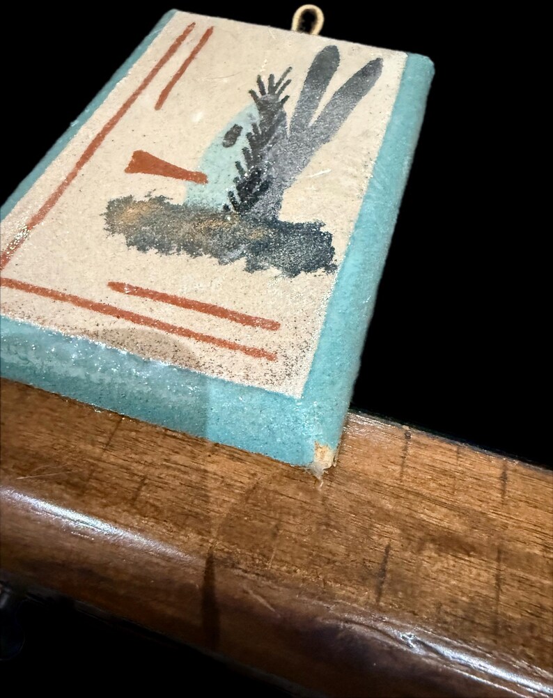 Op de afbeelding: Een rechthoekige wanddecoratie met een lichtblauwe rand en een beige midden. Het midden heeft een abstract ontwerp met bruine, zwarte en blauwe vormen. Er is een kleine goudkleurige haak aan de bovenkant bevestigd om op te hangen.