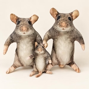 Può includere: Tre peluche a forma di topo di diverse dimensioni, caratterizzati da un realistico motivo di pelliccia grigia e bianca, grandi orecchie arrotondate, occhi scuri e un piccolo naso rosa. Le figure sono in piedi con le braccia distese sui lati.