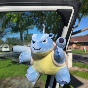 May include: A plush blue and yellow Blastoise toy with a suction cup attached. The toy is hanging from a car window. The Blastoise has two cannons on its back and is in a playful pose. The background shows a residential area with trees and houses.