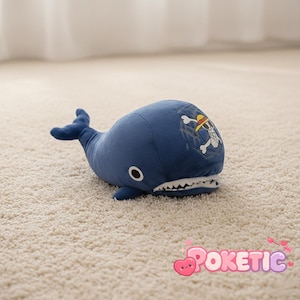 May include: A plush, blue whale toy with a white-toothed mouth and a tail fin. The whale has a pirate-themed skull and crossbones design on its back. The toy is lying on a beige carpet.