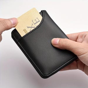 May include: A black leather cardholder with a card partially inserted. The card is tan with the letters "XE" visible. The cardholder is rectangular with a curved top edge and visible stitching.