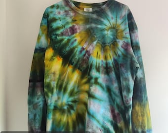 Può includere: Una maglia a maniche lunghe con motivo tie-dye nei toni del verde acqua, giallo, verde oliva e viola scuro. Il capo presenta un motivo a spirale ed è etichettato come XL Comfort Colors. È appesa a una gruccia di plastica nera.