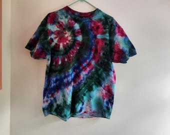 Può includere: Una t-shirt a maniche corte con girocollo caratterizzata da un motivo tie-dye nei toni del verde scuro, magenta e blu profondo. Il design include un evidente motivo a spirale sul lato superiore sinistro, con colori che si fondono insieme.