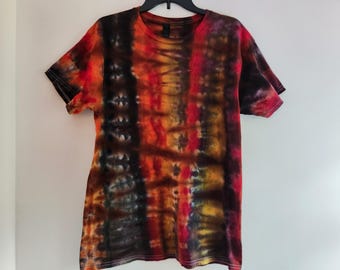 Può includere: Una t-shirt a maniche corte con girocollo caratterizzata da un motivo tie-dye con bande verticali di nero, marrone scuro, arancione brillante, giallo e rosso intenso. Il capo ha una vestibilità standard con scollo rotondo e maniche orlate, mostrando un gradiente di colore coerente sulla superficie anteriore.