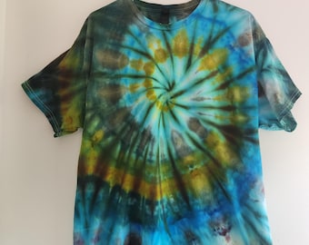 Può includere: Una t-shirt a maniche corte con un motivo tie-dye a spirale nei toni del blu brillante, verde bosco, giallo senape e nero intenso. Il capo presenta uno scollo rotondo ed è appeso a una gruccia in plastica di colore azzurro.