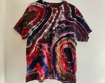 Può includere: Una t-shirt a maniche corte con un motivo tie-dye a geode nei toni del rosso scuro, nero, marrone e bianco. Il design è composto da forme circolari organiche e concentriche che imitano l'aspetto di una pietra preziosa tagliata.