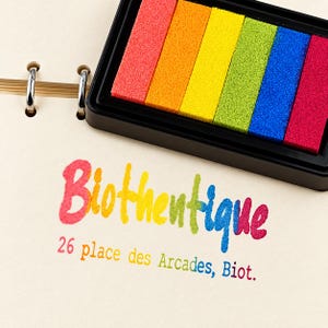 May include: A multi-colored ink pad with six rectangular segments in red, orange, yellow, green, blue, and magenta. Below the ink pad, the text "Biothentique" and "26 place des Arcades, Biot." is printed in a rainbow-colored font on paper.