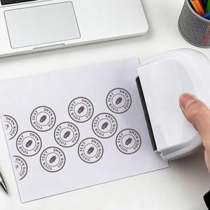 Op de afbeelding: Een witte stempel brengt cirkelvormige zwarte inktstempels aan op wit papier. De stempels hebben de tekst "YOUR NAME" en "TEXT" rond een bladontwerp. De stempel staat op een wit oppervlak.