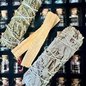 May include: An assortment of small glass jars with cork stoppers, each labeled with a different gemstone name. Two bundles of dried sage and two sticks of palo santo are also visible, arranged in a dark box.