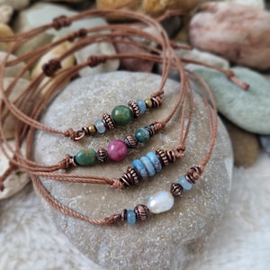 Könnte beinhalten: Vier verstellbare Armbänder aus dünner brauner Kordel, jedes mit einer einzigartigen Anordnung aus bunten Perlen und kupferfarbenen Metallzwischenstücken. Die Perlen umfassen verschiedene Grün-, Rosa- und Blautöne sowie eine weiße Perle.