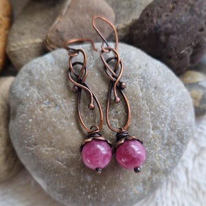 May include: A pair of dangle earrings featuring dark copper wire formed into an infinity-style loop design. Each earring is accented with a single round, polished magenta-colored gemstone bead secured at the base with wrapped copper wire.