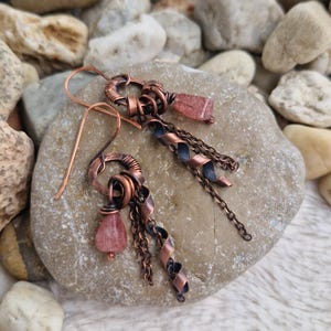 May include: A pair of dangle earrings crafted from oxidized copper wire, featuring coiled wire loops, delicate metal chains, and twisted copper ribbons. Each earring is accented with a small, translucent, pale pink strawberry quartz gemstone drop.