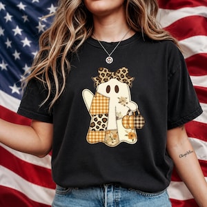 May include: A black short-sleeve t-shirt featuring a central graphic of a ghost character. The ghost is decorated with patchwork patterns including leopard print, orange plaid, and floral designs, wearing a leopard print bow and holding a pumpkin.