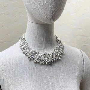 May include: A silver rhinestone necklace with a cluster of clear stones. The necklace is on a white mannequin.