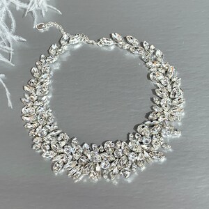 May include: A silver necklace with a delicate chain and a large, ornate design of clear crystal stones. The stones are arranged in a cluster of leaves, creating a flowing, organic look.