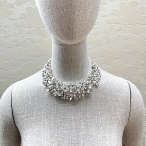 May include: A silver statement necklace with a cluster of clear and pearlescent crystals. The necklace is on a white mannequin.