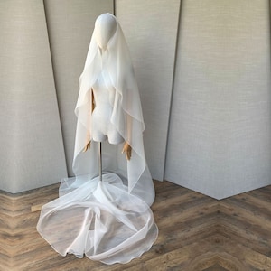 Pu&ograve; includere: Un velo da sposa in tulle bianco con un lungo strascico fluido. Il velo &egrave; drappeggiato su un manichino ed &egrave; esposto su uno sfondo grigio chiaro.
