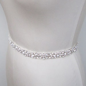 May include: A white satin bridal sash with a crystal embellishment. The sash is shown on a white dress form.