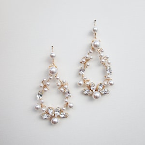 May include: A pair of gold dangle earrings with a teardrop shape. The earrings are adorned with small white pearls and clear rhinestones.