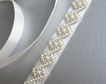 May include: A white satin ribbon with a beaded embellishment. The embellishment is made of small white pearls and clear rhinestones arranged in a geometric pattern.
