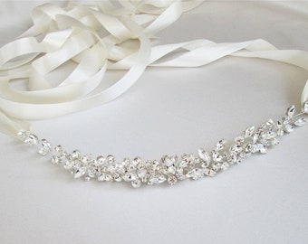May include: A white satin ribbon sash with a crystal embellishment. The embellishment features a cluster of clear rhinestones in a floral design.