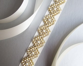 May include: A white satin ribbon with a gold and pearl embellishment. The embellishment is a repeating pattern of diamond shapes with pearls and rhinestones.