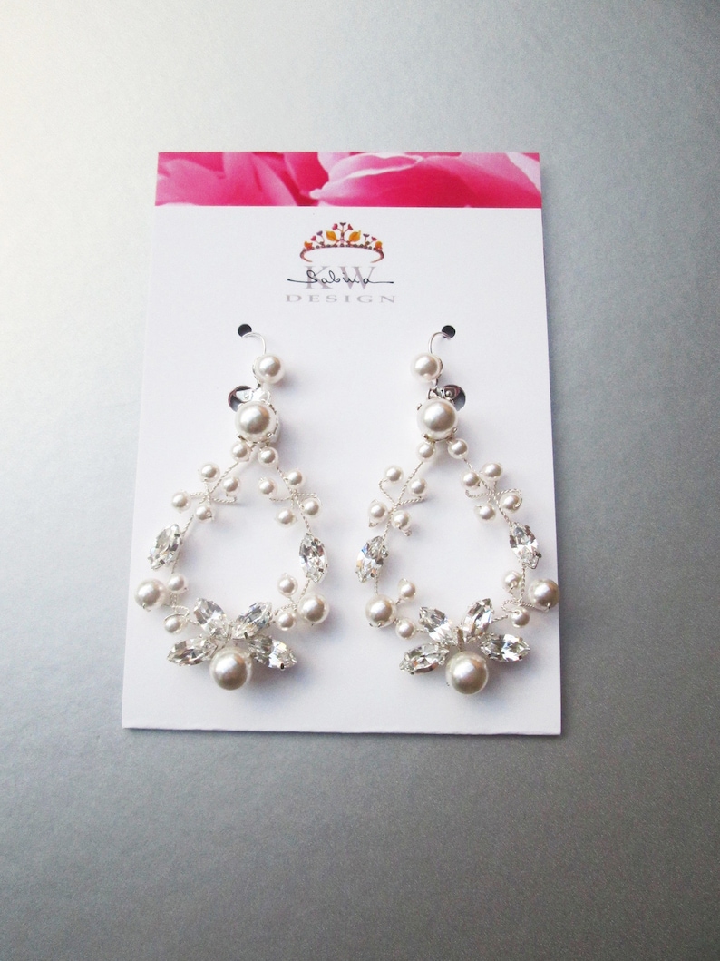 May include: A pair of silver dangle earrings with a floral design featuring clear crystals and white pearls. The earrings are displayed on a white card with the text "KW DESIGN" printed on it.