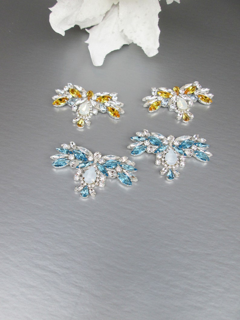 May include: Four decorative embellishments with crystal-like stones. Two are blue and two are yellow.