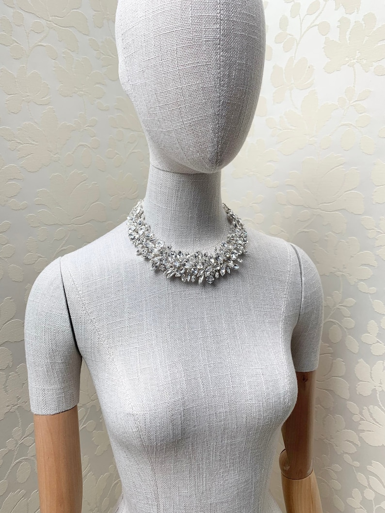 May include: A silver rhinestone necklace with a cluster of clear stones. The necklace is on a white mannequin.