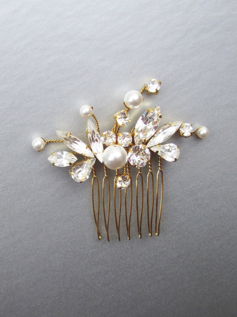 Puede incluir: Un peine de pelo dorado con un grupo de piedras de cristal transparentes y perlas blancas. El peine tiene un dise&ntilde;o floral delicado.