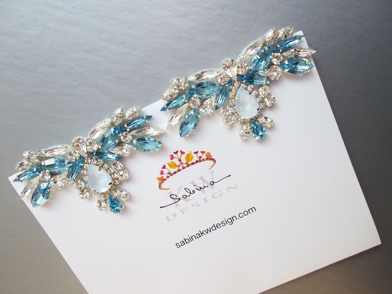 May include: A pair of large, statement earrings featuring a cluster of blue and clear rhinestones. The earrings are arranged in a cascading design, with the larger stones at the top and the smaller stones at the bottom. The earrings are set on a silver base.
