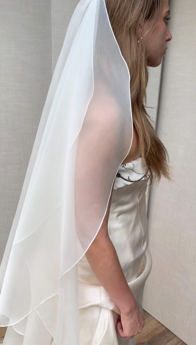 Pu&ograve; includere: Un velo da sposa bianco a un solo livello con un bordo a festoni. Il velo &egrave; realizzato in tessuto morbido e trasparente ed &egrave; drappeggiato sulla testa e sulle spalle della sposa.